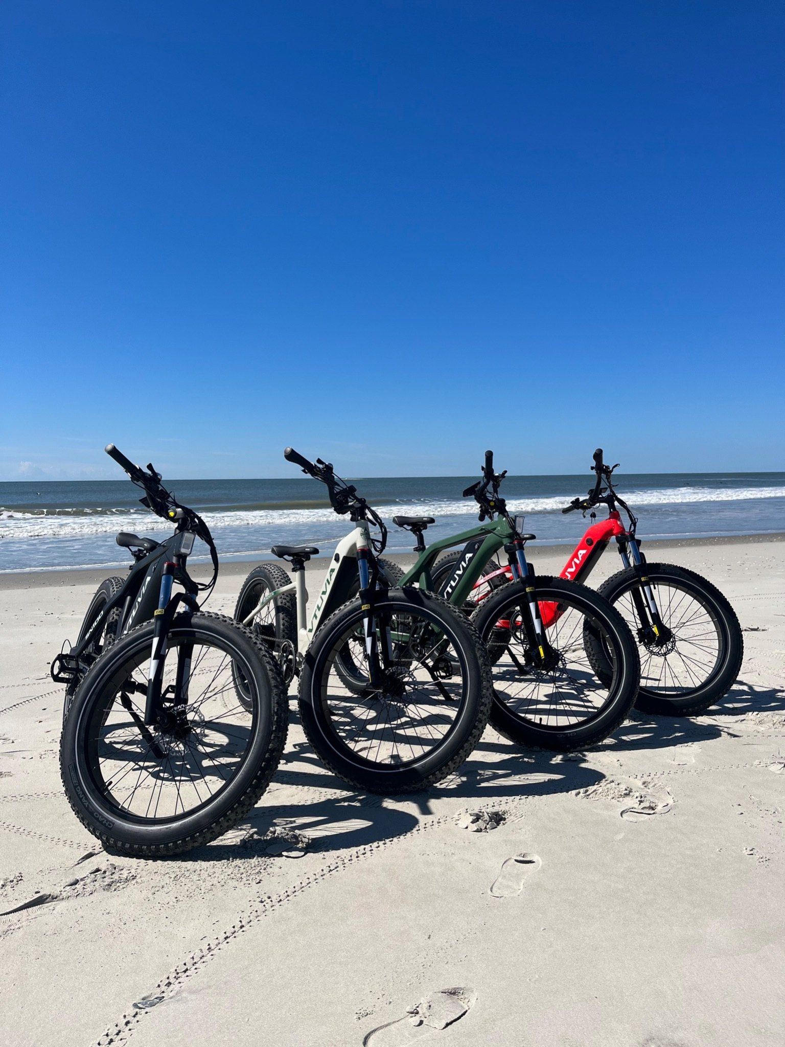 Four fat-tire step-through e-bikes in black, cream, green and red parked side by side on the sand, ocean behind them