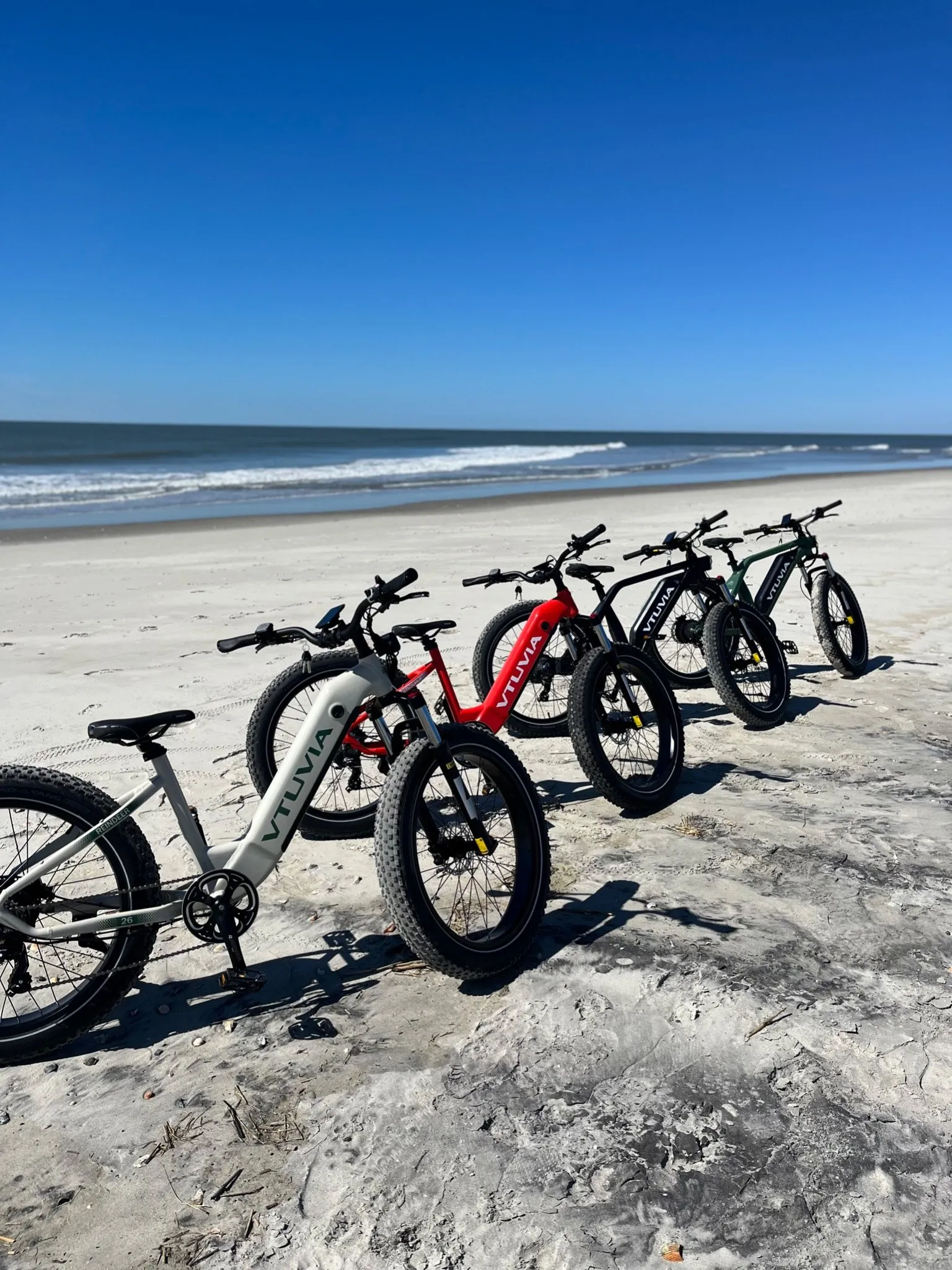A row of fat-tire e-bikes on their kickstands on the sand, a cream bike in front